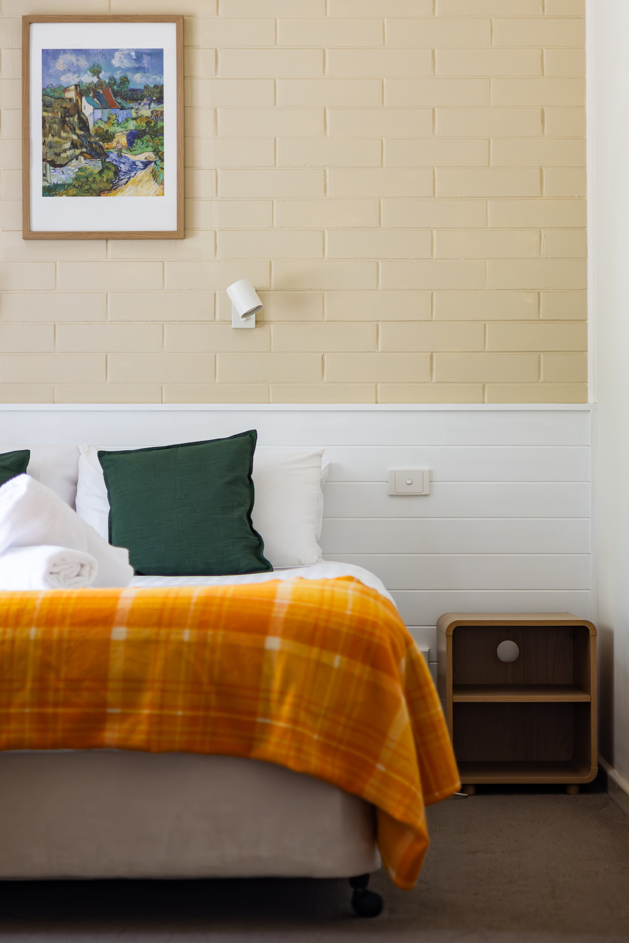 A made-up queen bed with green cushions and an orange checked throw, a framed print above