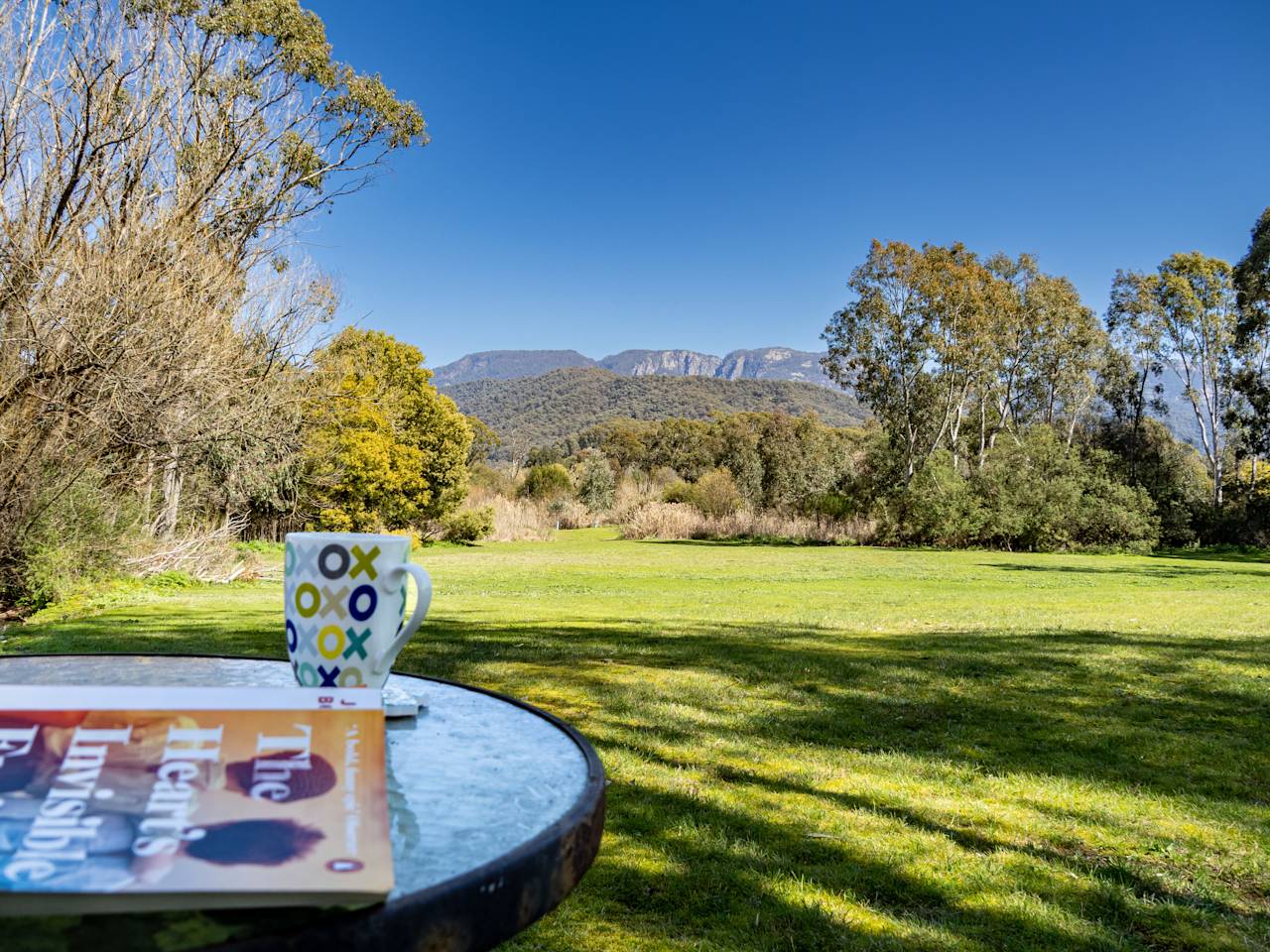 A coffee mug and a book on a round outdoor table, with the lawn and the ranges beyond