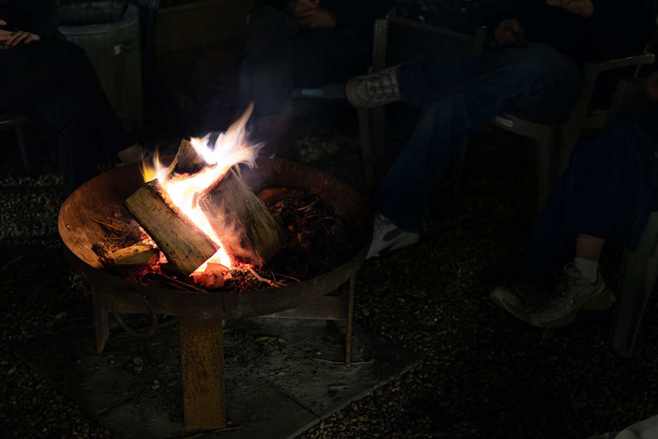 Logs burning in the fire pit at night, with guests in camp chairs around it