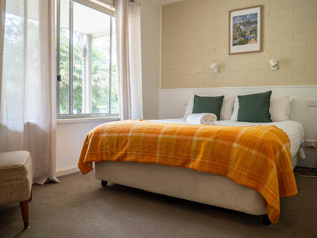 A queen bedroom with an orange check blanket and green cushions, lit by a window onto the garden