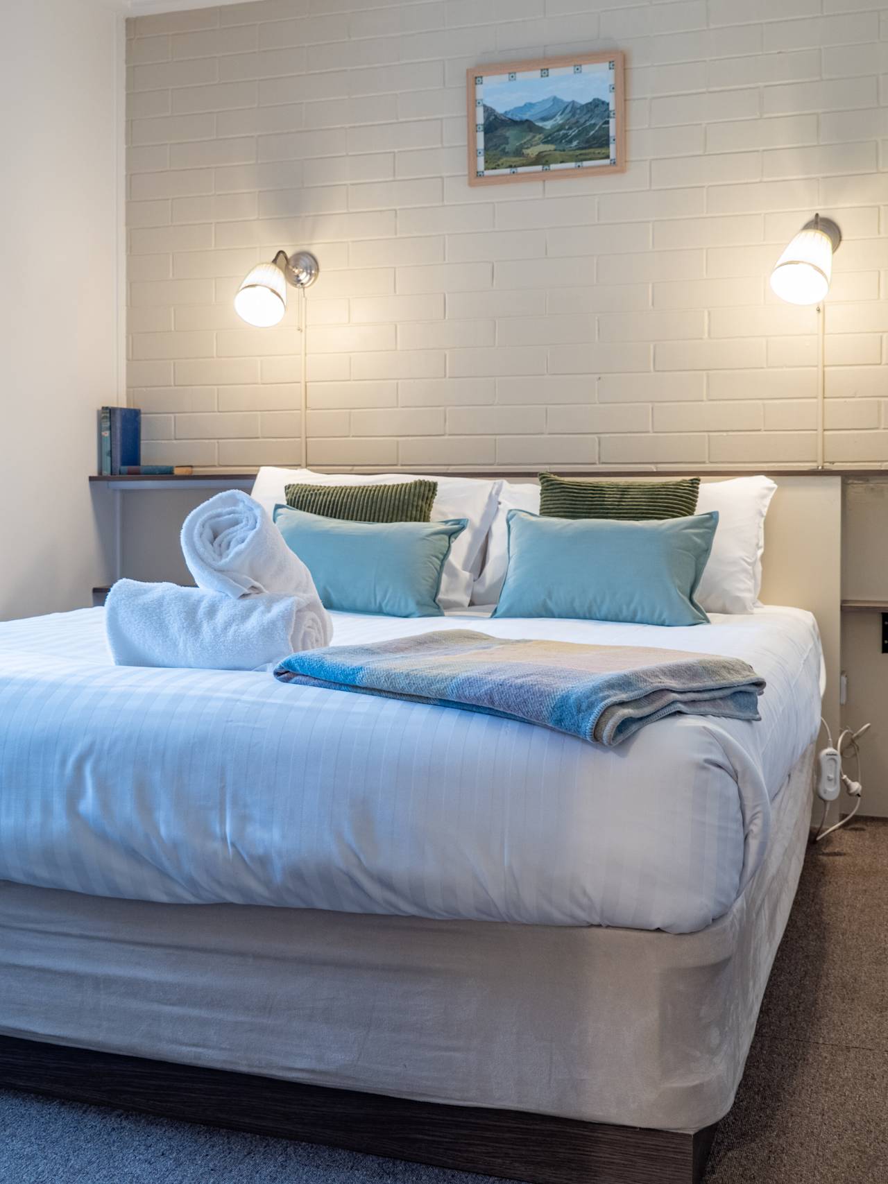 A made-up queen bed with blue cushions and a folded towel, under two wall lamps and a small mountain painting