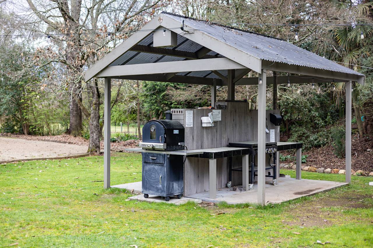 The covered barbecue shelter on the lawn, with a hooded barbecue and a bench