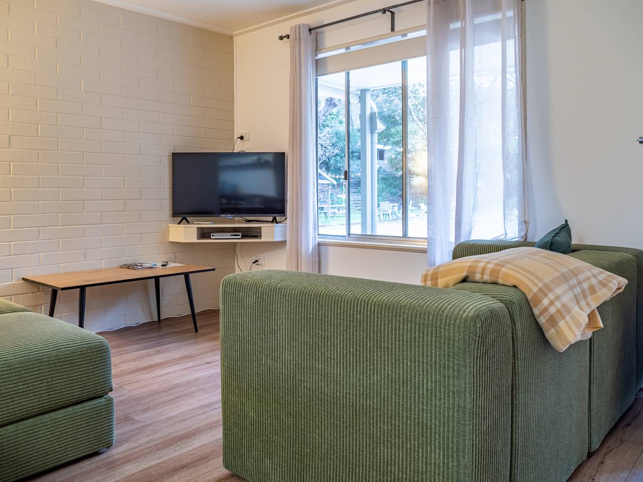 A living room with a green corduroy sofa, a low timber table and a television beside a bright window