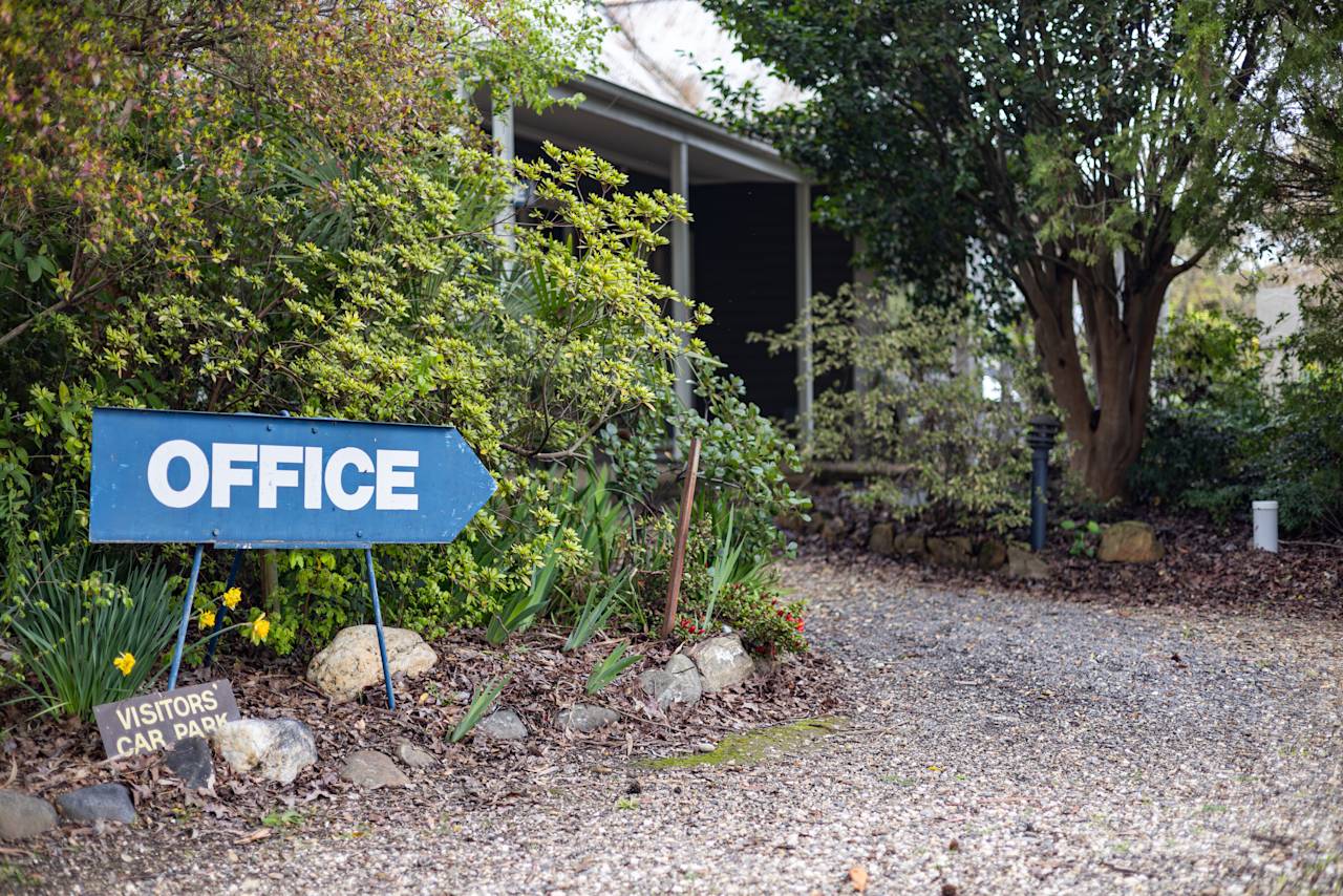 A blue Office sign pointing along the gravel drive, past garden beds and the units